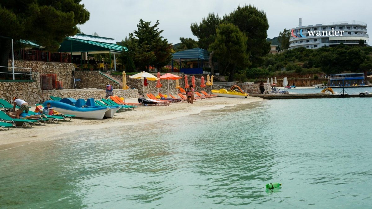 Empty beach with colorful lounge chairs and umbrellas, paddle boats, and a few people near the water. Trees and a modern building are visible in the background.