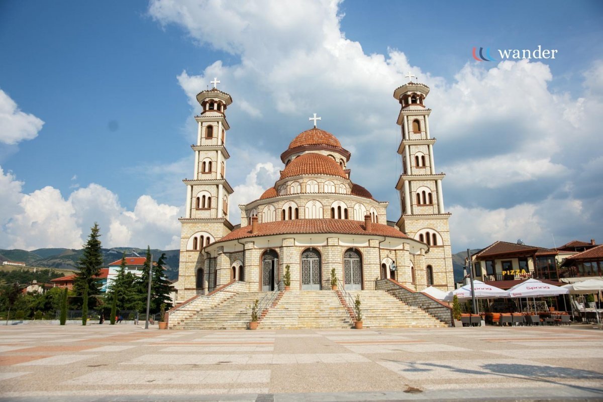 A large church with multiple domes and two tall bell towers, situated in an open square with a few trees and outdoor seating nearby, under a partly cloudy sky.