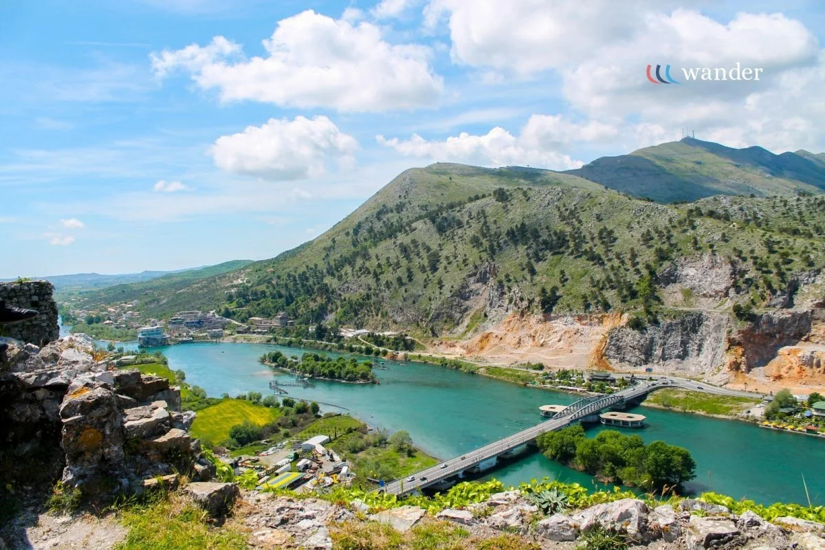 Scenic view of a river running through a valley with mountains in the background, a bridge crossing the river, and a partly cloudy sky.