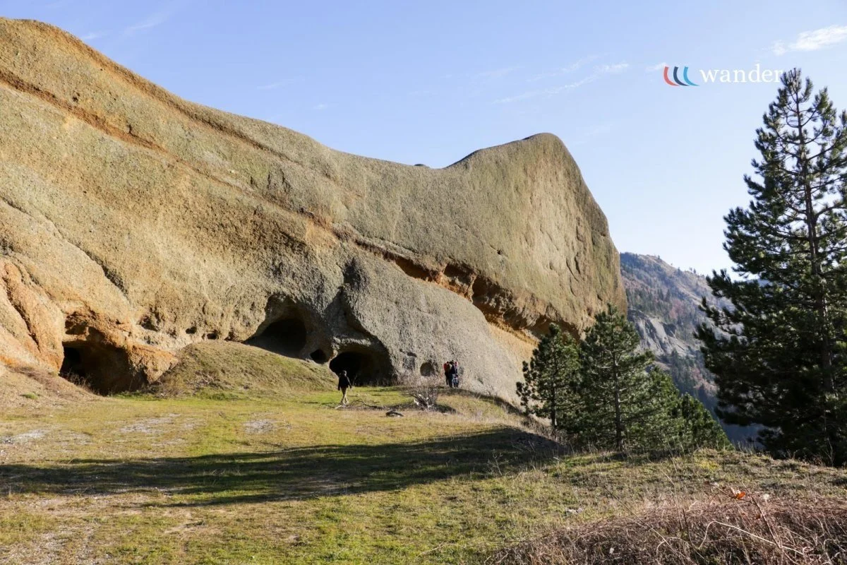 People standing near cave openings in a large rock formation with grassy ground and pine trees, mountain scenery in the background, under a clear sky.