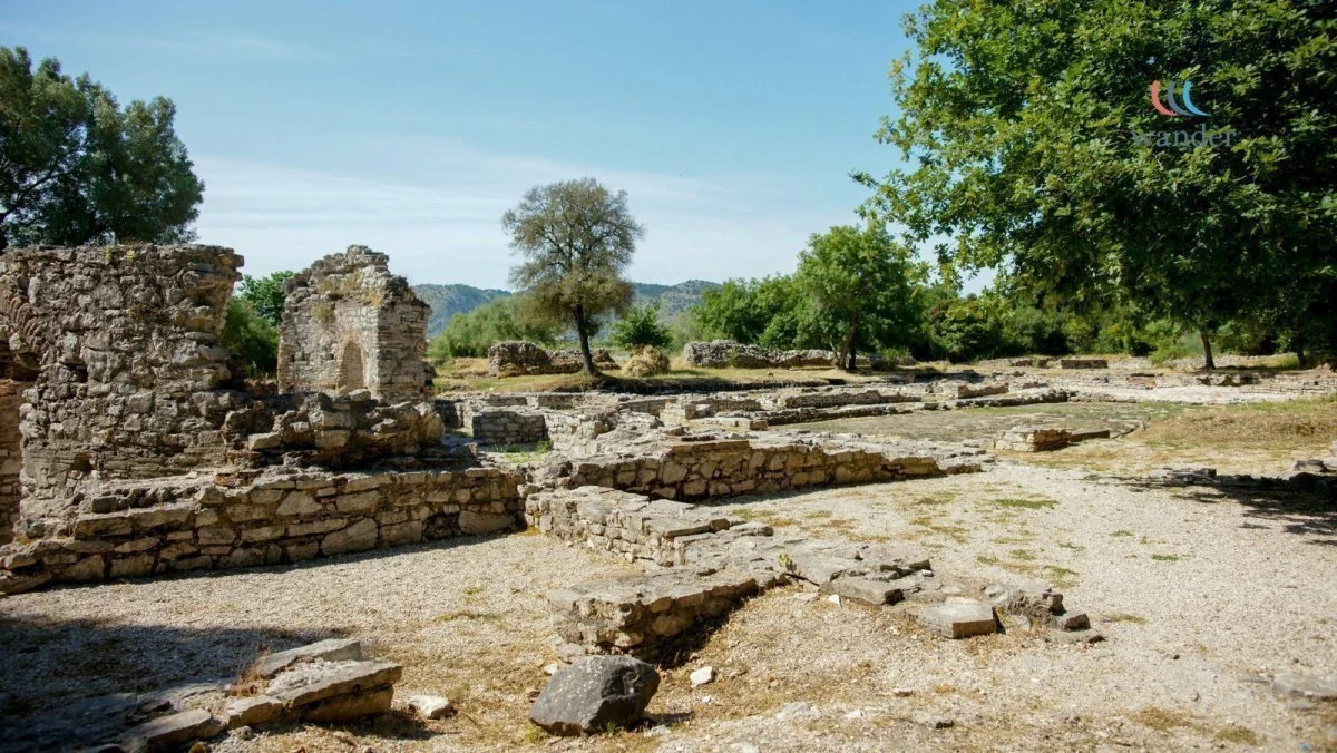 Ruin of an ancient archaeological site with stone foundations and partial walls, surrounded by trees and a clear blue sky.