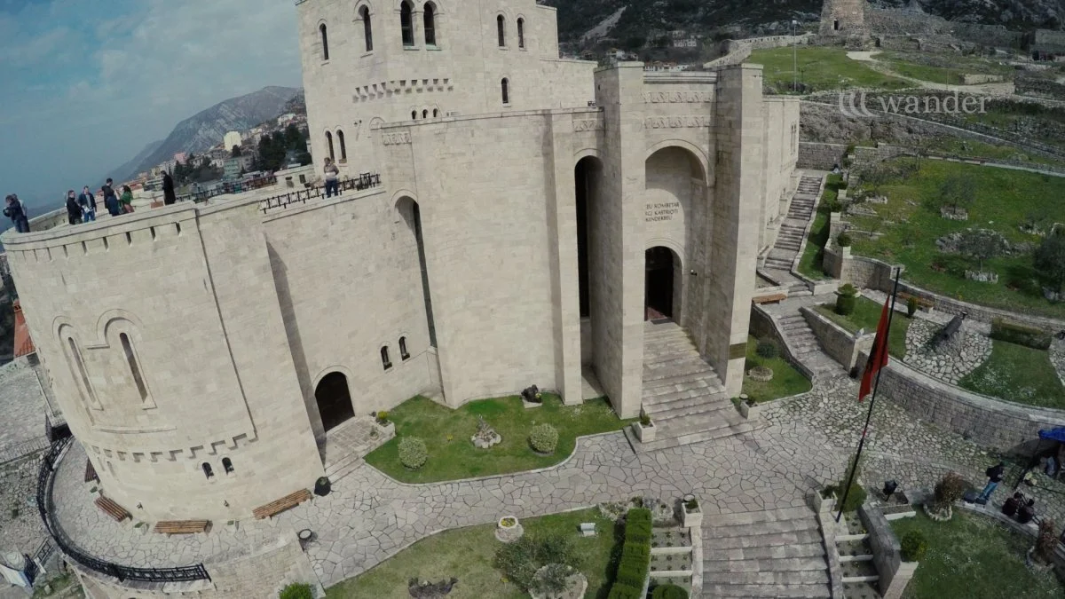 A stone fortress with a curved wall and an entrance, surrounded by a paved courtyard with benches and greenery, overlooking mountains and a town in the background.
