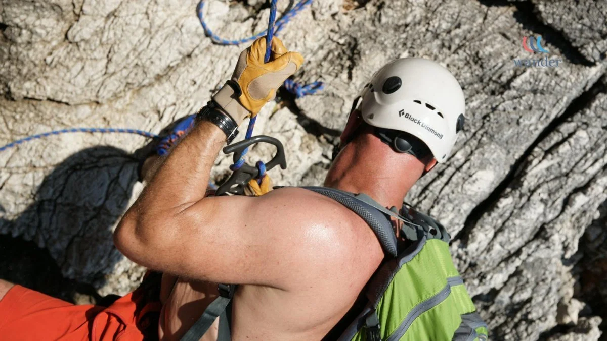 A man rock climbing outdoors, wearing a white helmet, a green backpack, and a sleeveless shirt, is secured with climbing gear and ropes on a rocky cliff face.