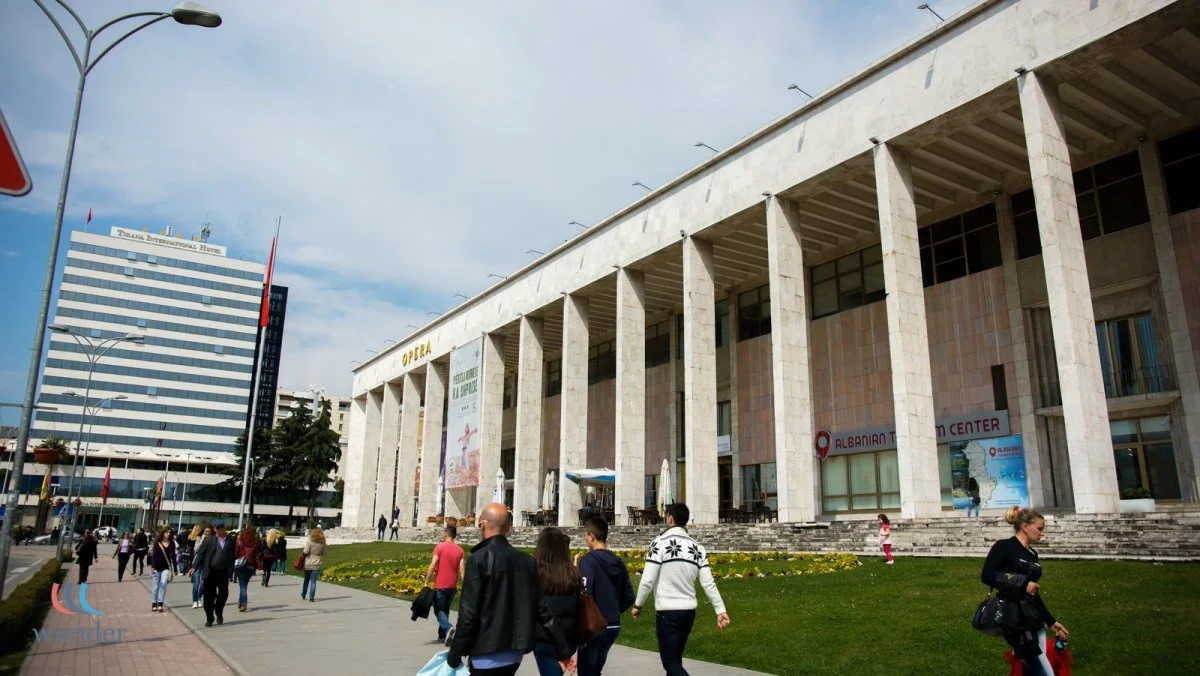 People walking near a large building with tall columns, on a city street with a grassy area, and a tall modern building in the background.