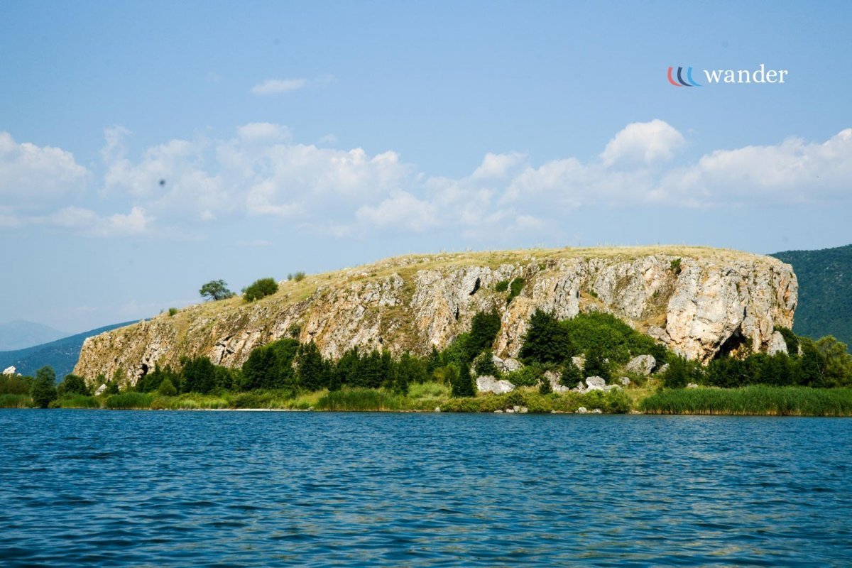 A large rocky island surrounded by blue water with green trees and grass, under a partly cloudy sky. The word 'wander' appears in the top right corner.