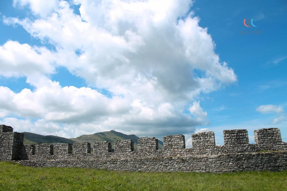 Ancient stone wall with battlements in a grassy field under a blue sky with clouds, mountains in the background.