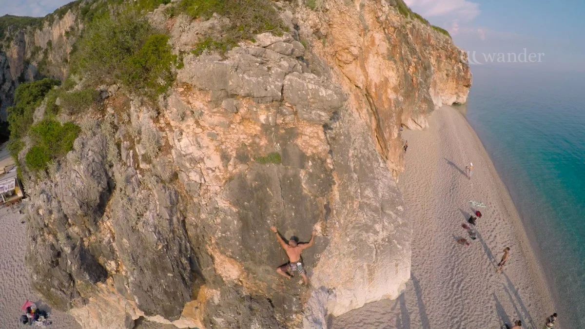 A person rock climbing on a steep cliff with a sandy beach and ocean below, and several people on the beach.