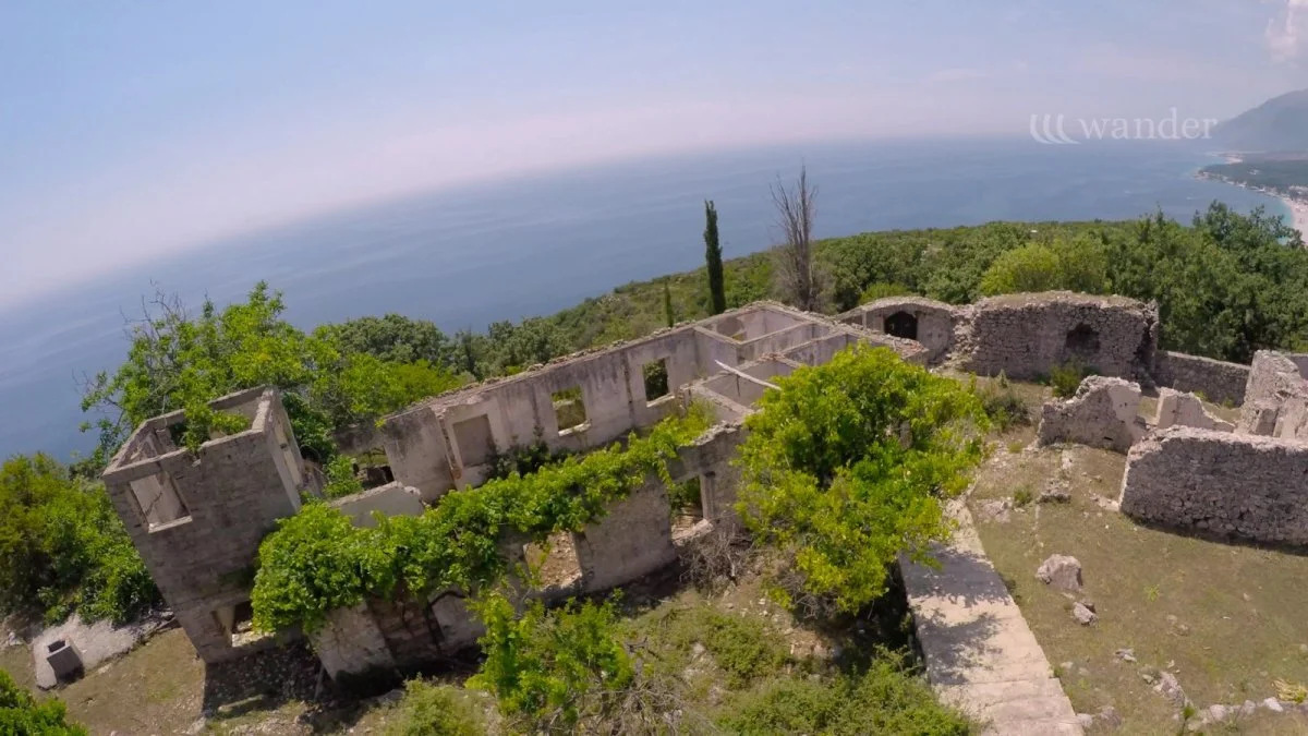 Aerial view of ruins of an old stone building with multiple rooms and open windows, surrounded by green trees, overlooking a body of water and mountains in the distance.