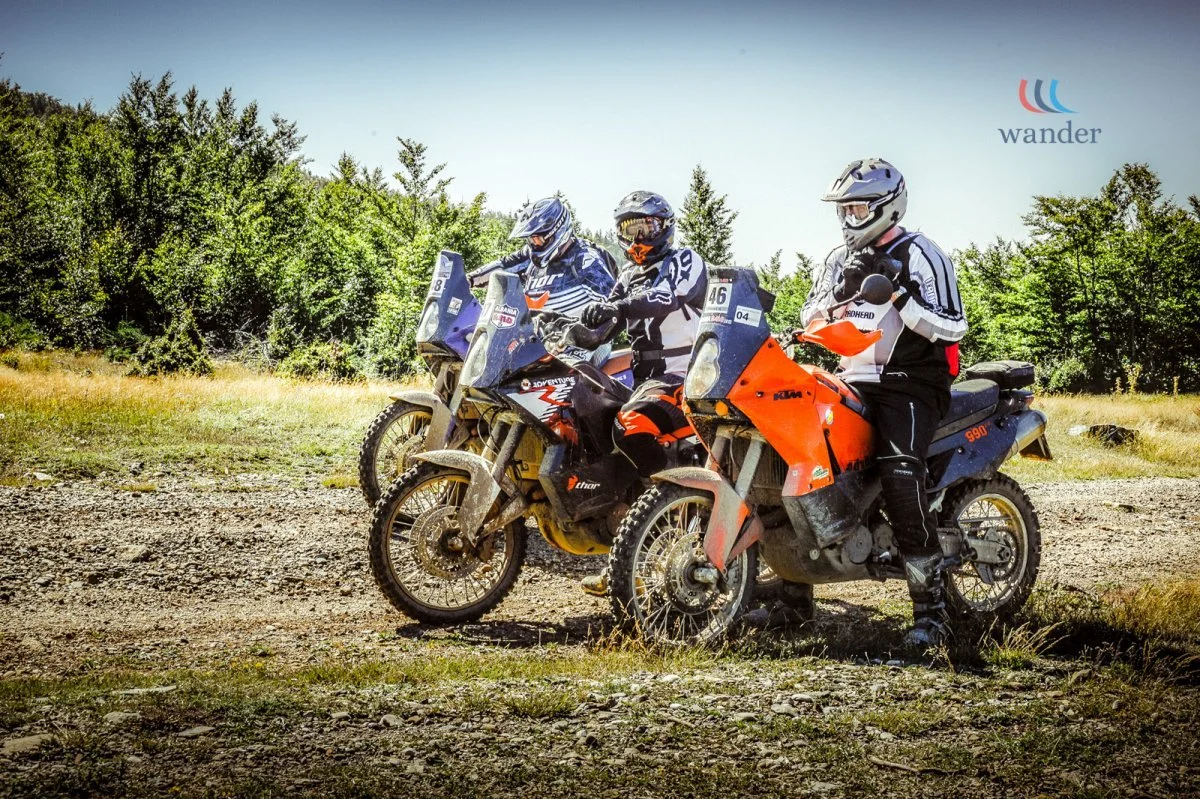 Three individuals in riding gear on adventure motorcycles, sitting on dirt bikes in an outdoor setting with trees and a dirt path.