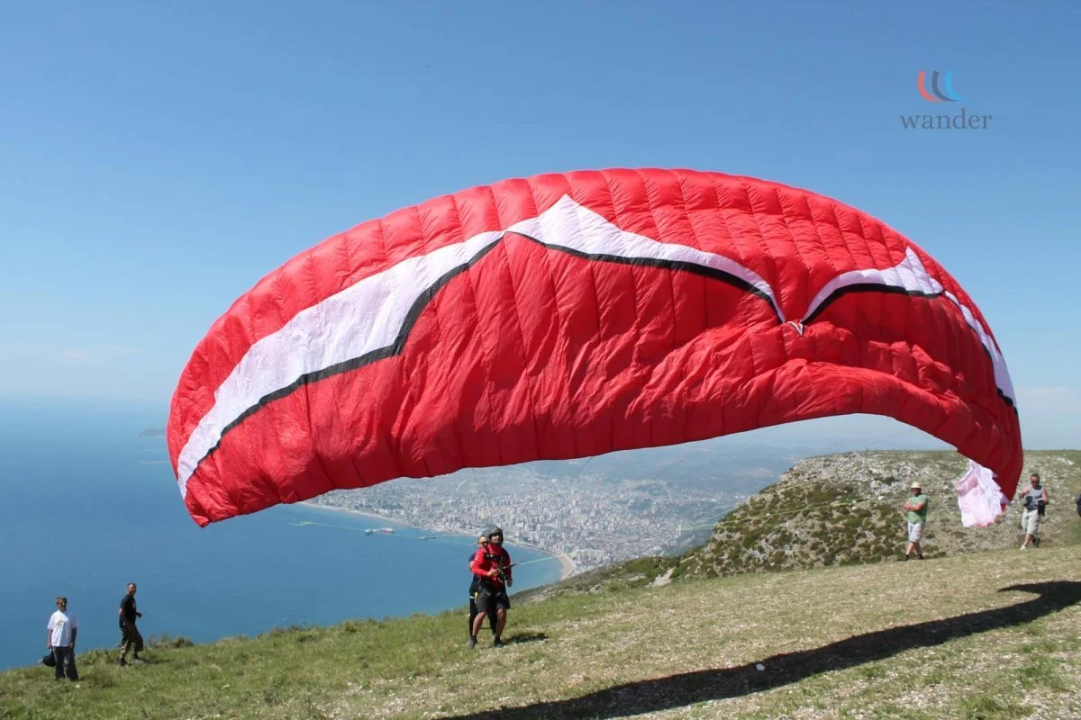 People preparing for paragliding on a grassy hillside near a body of water, with a large red and white parachute partially inflated and in mid-flight.