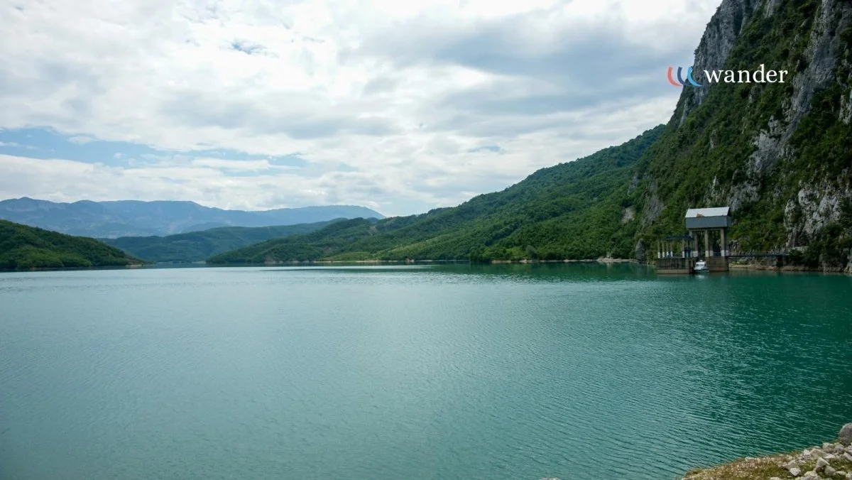 Scenic landscape of a calm lake surrounded by green mountains and clouds in the sky, with a small pavilion on the right side beside the water.