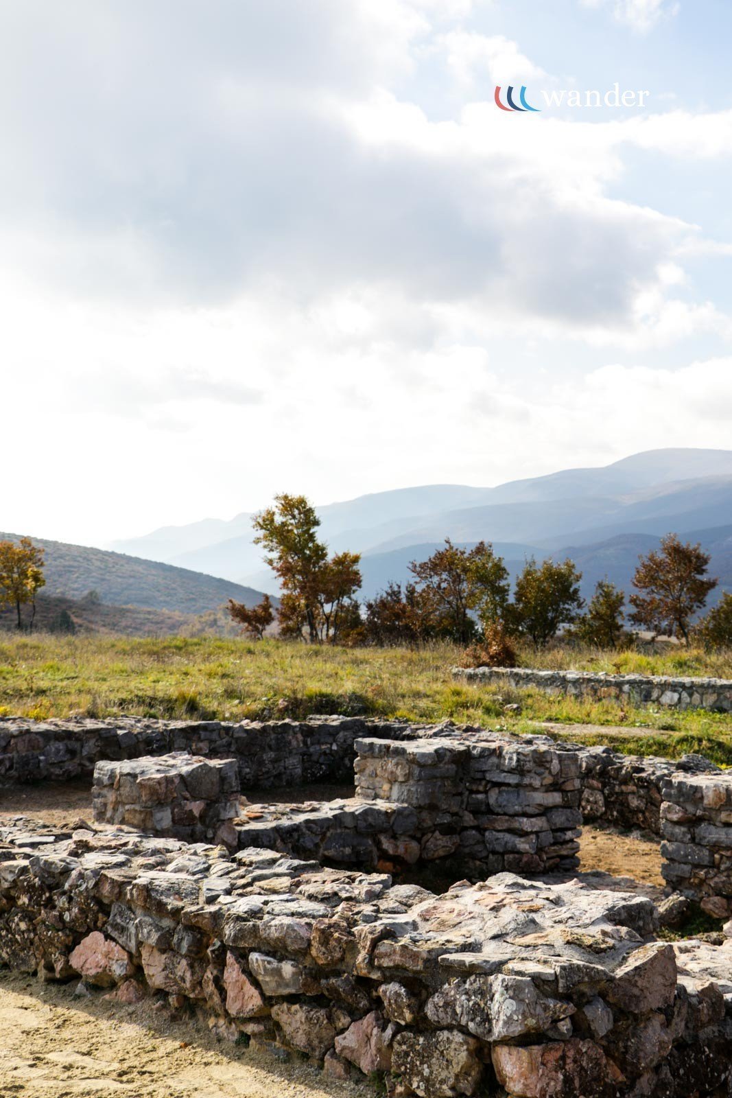 Ruins of ancient stone structures with a mountainous landscape and trees in the background under a partly cloudy sky.