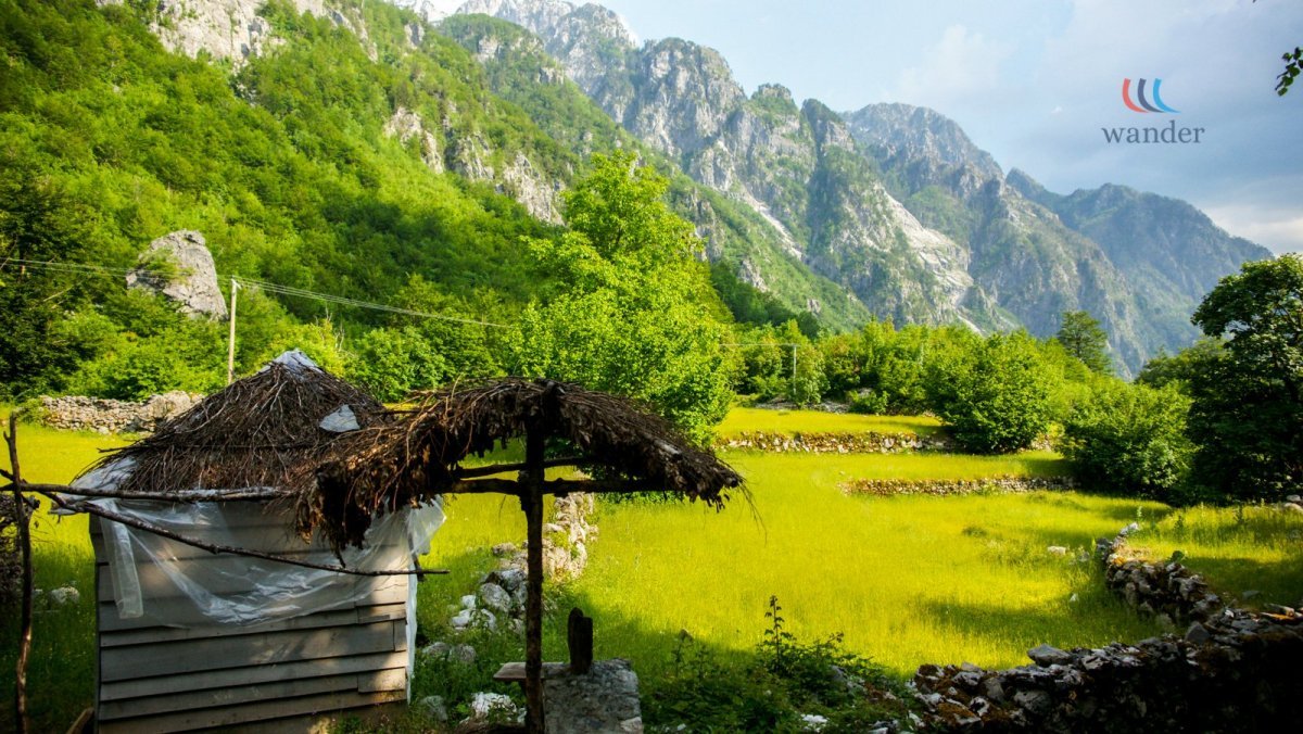 A lush green rural landscape with mountains in the background, a small rustic hut with a thatched roof, and stone walls surrounding the fields.