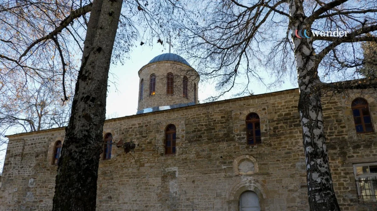 A stone church with arched windows seen through tree branches, with a domed roof and a cross on top, against a blue sky.