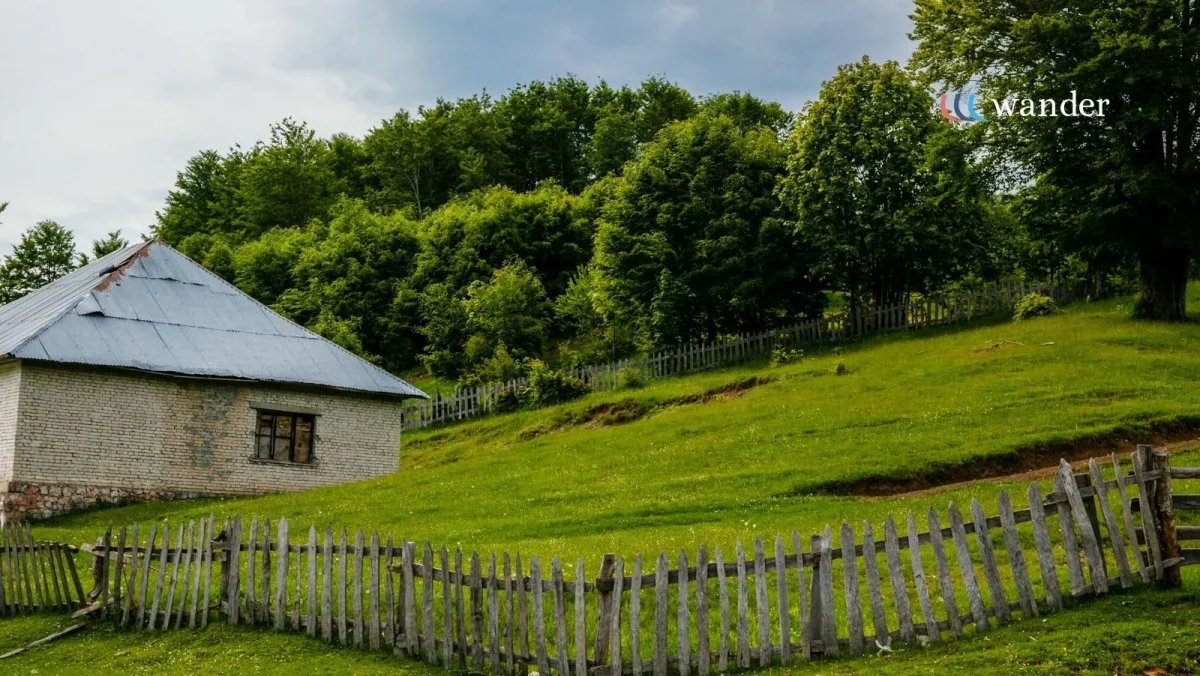 A rural landscape with a small brick house with a metal roof, green grass, a wooden fence, and trees on a hillside under a cloudy sky.