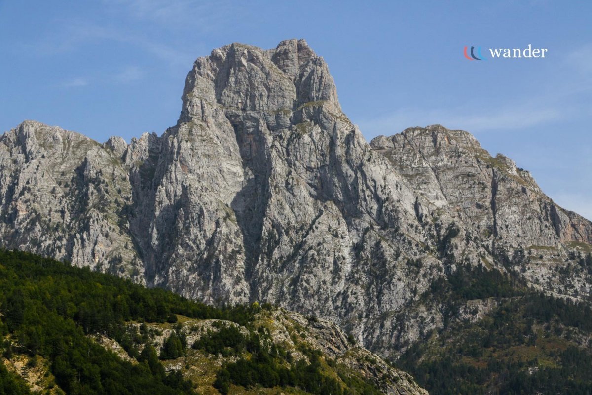 A large rocky mountain with steep cliffs and lesser green forested areas at the base, under a light blue sky with few clouds.