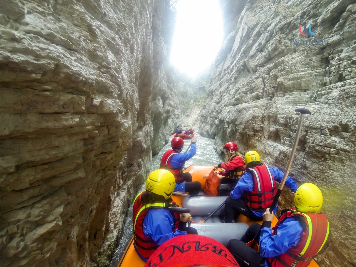 Group of people whitewater rafting in a narrow canyon with high rock walls, wearing helmets and life jackets.