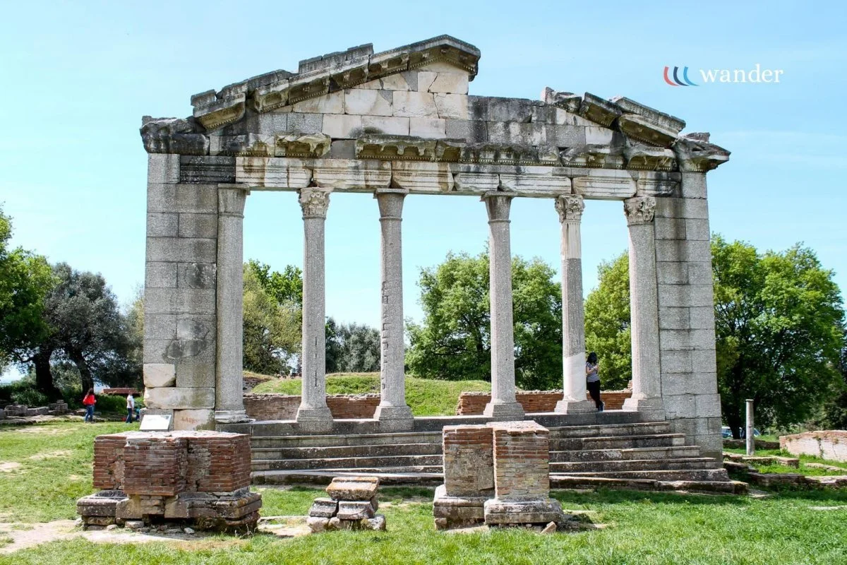 Ancient Greco-Roman temple ruins with columns and stairs, surrounded by grass and trees, under a clear sky.