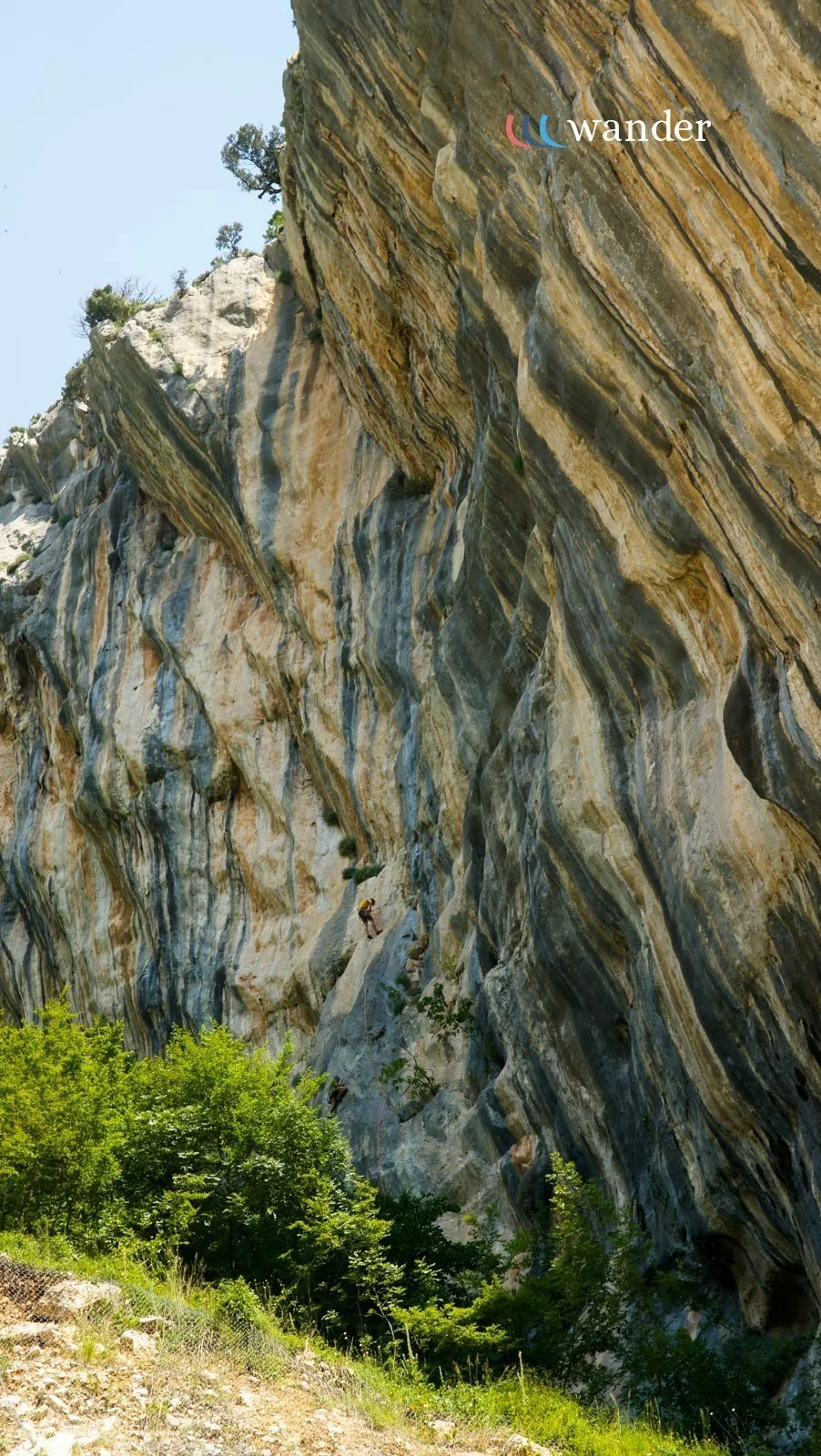 A large, colorful cliff face with layered stripes of various shades of gray, brown, and tan, with a person rock climbing in the lower part of the image, and green bushes at the base.