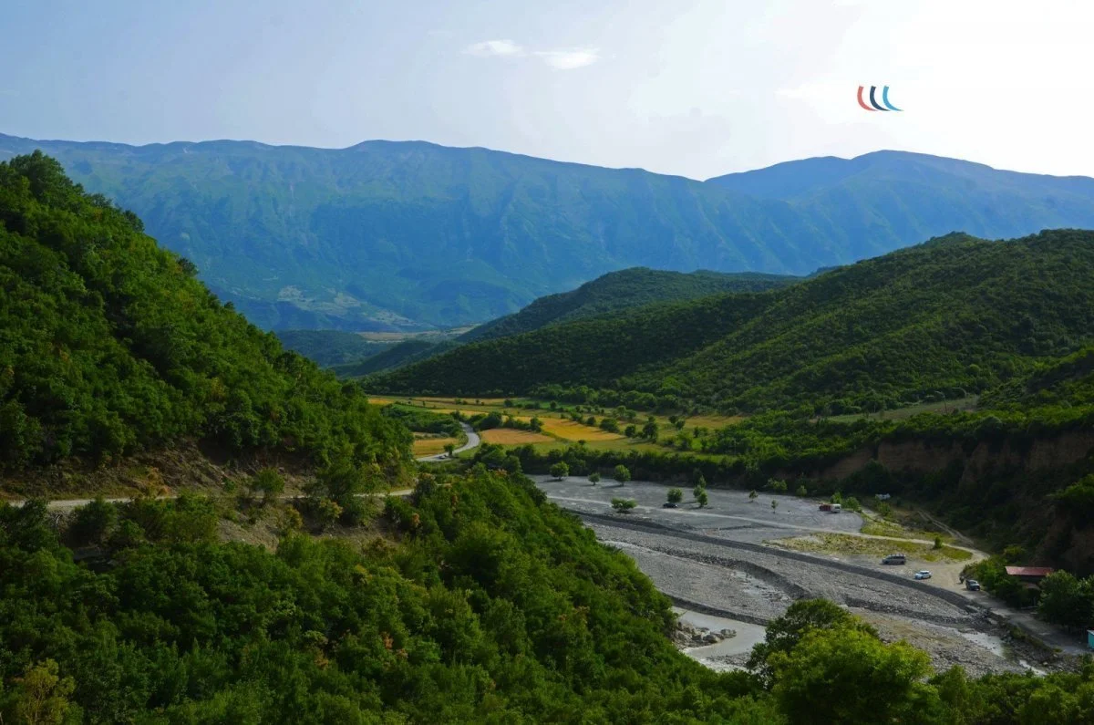 A scenic view of a valley with green hills and mountains, a winding river, and a few cars parked near the riverbank.