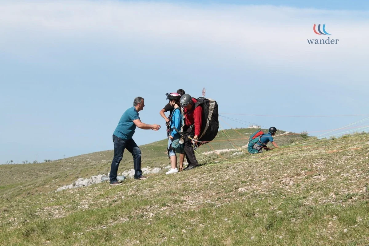 People helping a young person with parachuting gear as they prepare for tandem jump. Others are on the ground adjusting equipment, on a grassy field with a hill and sky in the background.