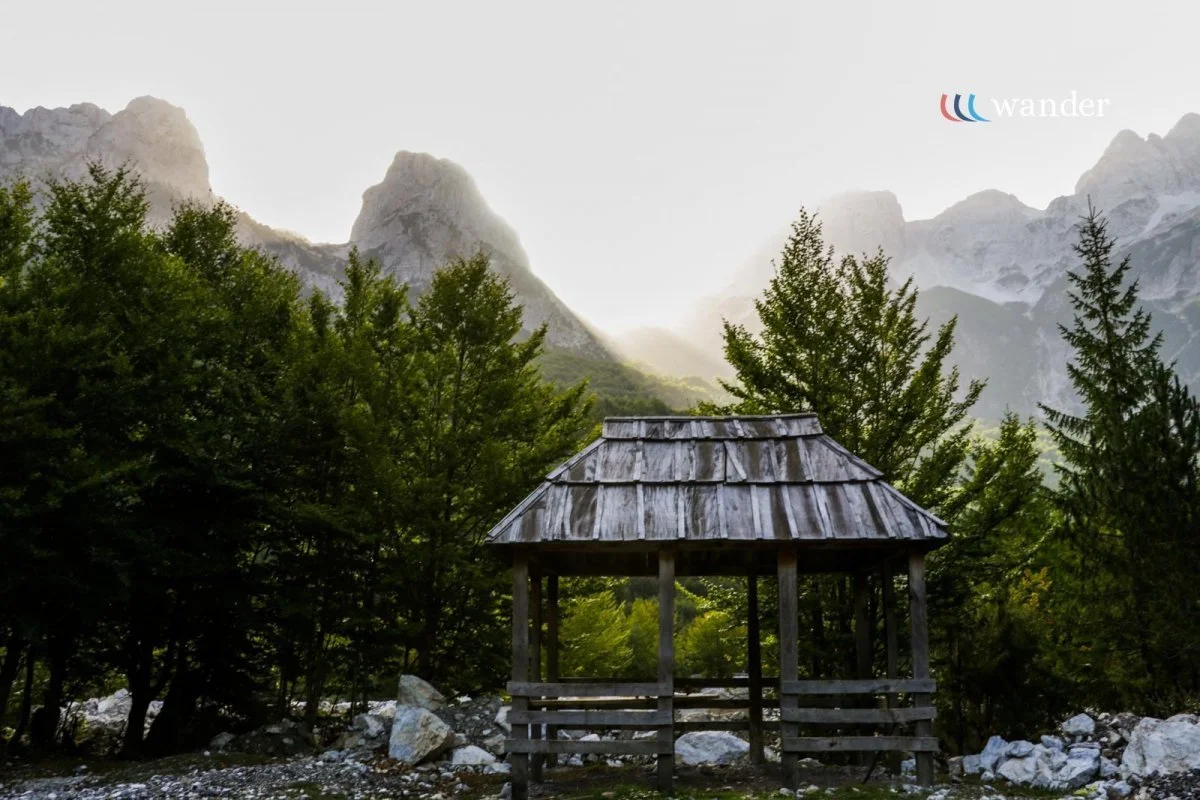 A rustic wooden pavilion with a shingled roof, surrounded by green trees and mountains in the background.