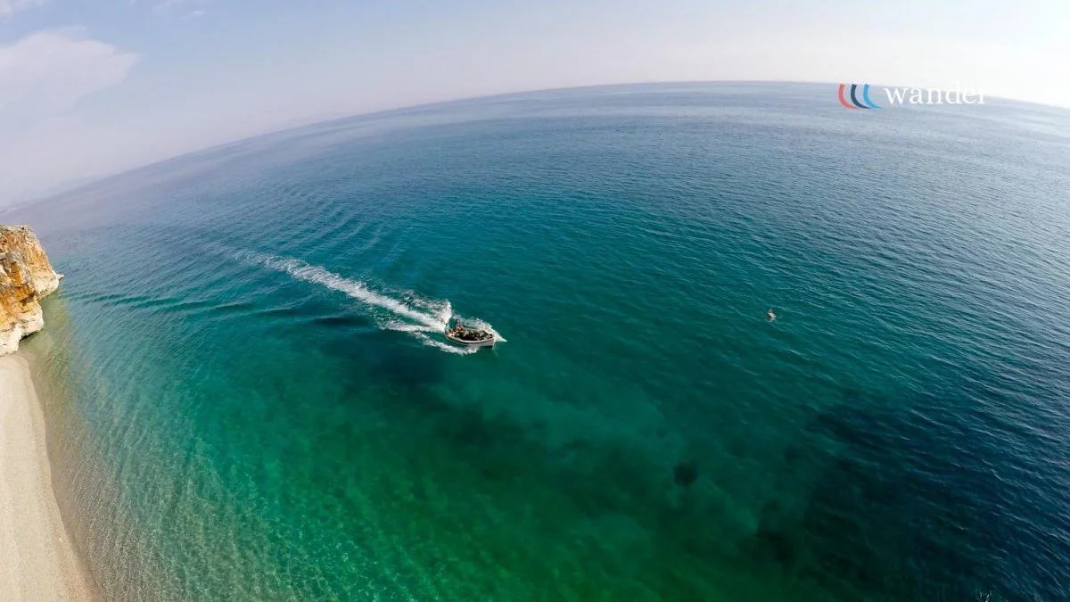 A boat sailing in clear blue-green ocean water near a sandy beach and rocky cliffs, with a small buoy floating nearby.