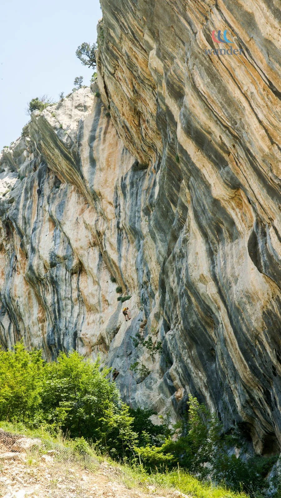 A person rock climbing on a large, colorful, striped cliff face with trees and bushes at the base, blue sky overhead.