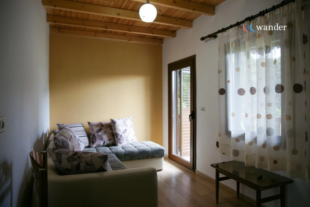 Living room with beige wall, white sofa with floral and striped pillows, wooden ceiling, window with polka dot curtains, door leading outside, small wooden table, and ceiling light