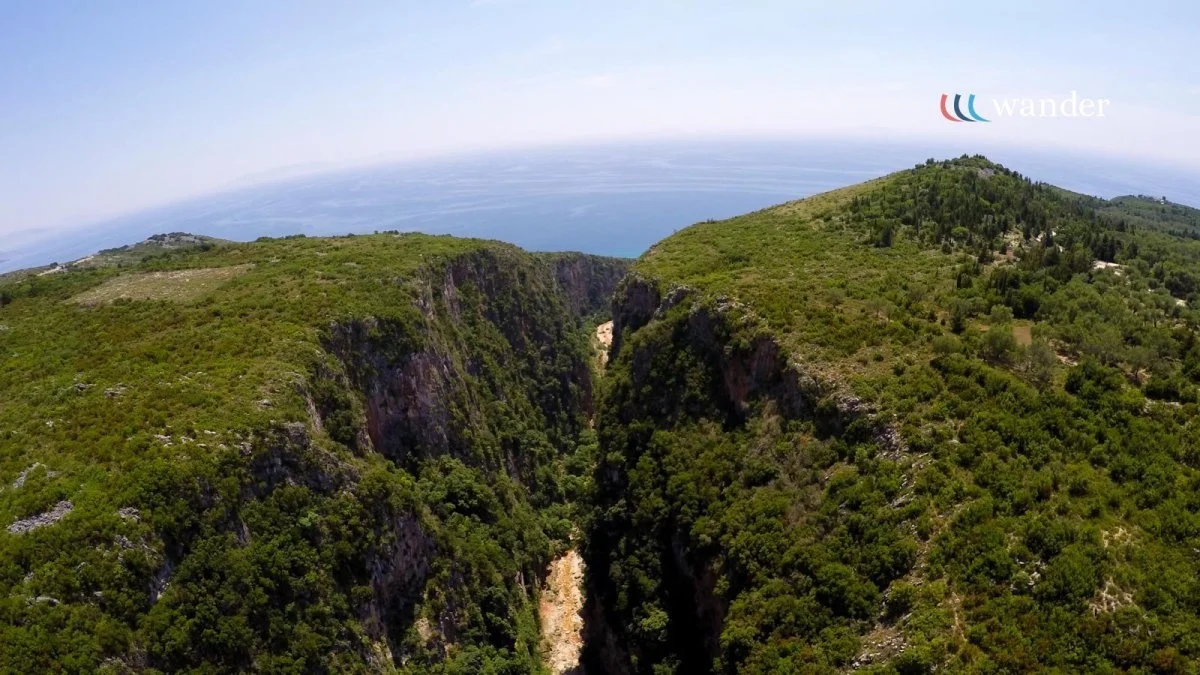 Aerial view of a deep canyon with rocky cliffs and dense green vegetation, overlooking the ocean in the background under a clear blue sky.