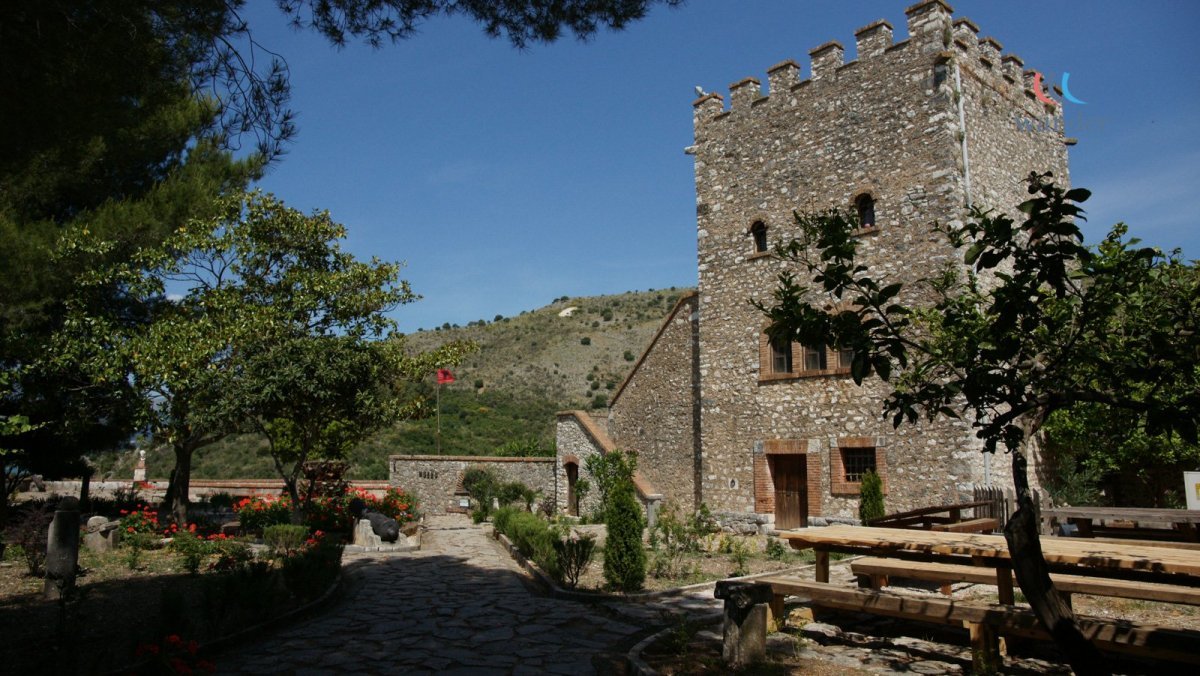 A stone castle tower with battlements, surrounded by green trees and a garden with flowers, in a mountainous area under a clear blue sky.