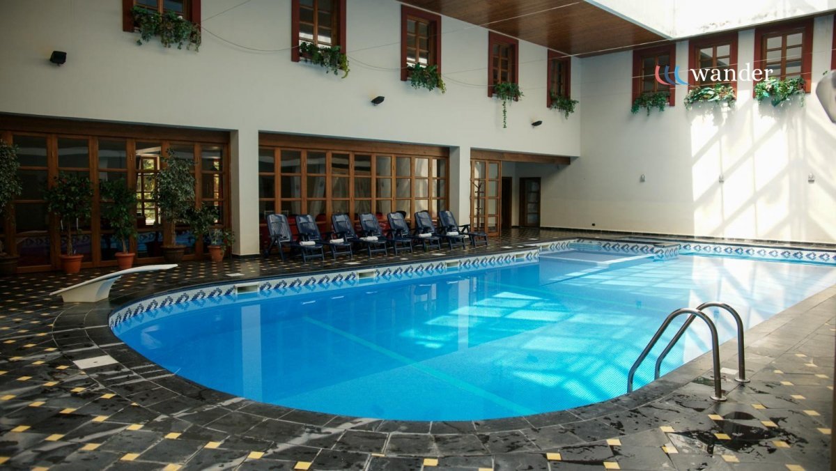 Indoor swimming pool with blue water, black and yellow tiled flooring, lounge chairs, potted plants, and large windows with wooden frames.
