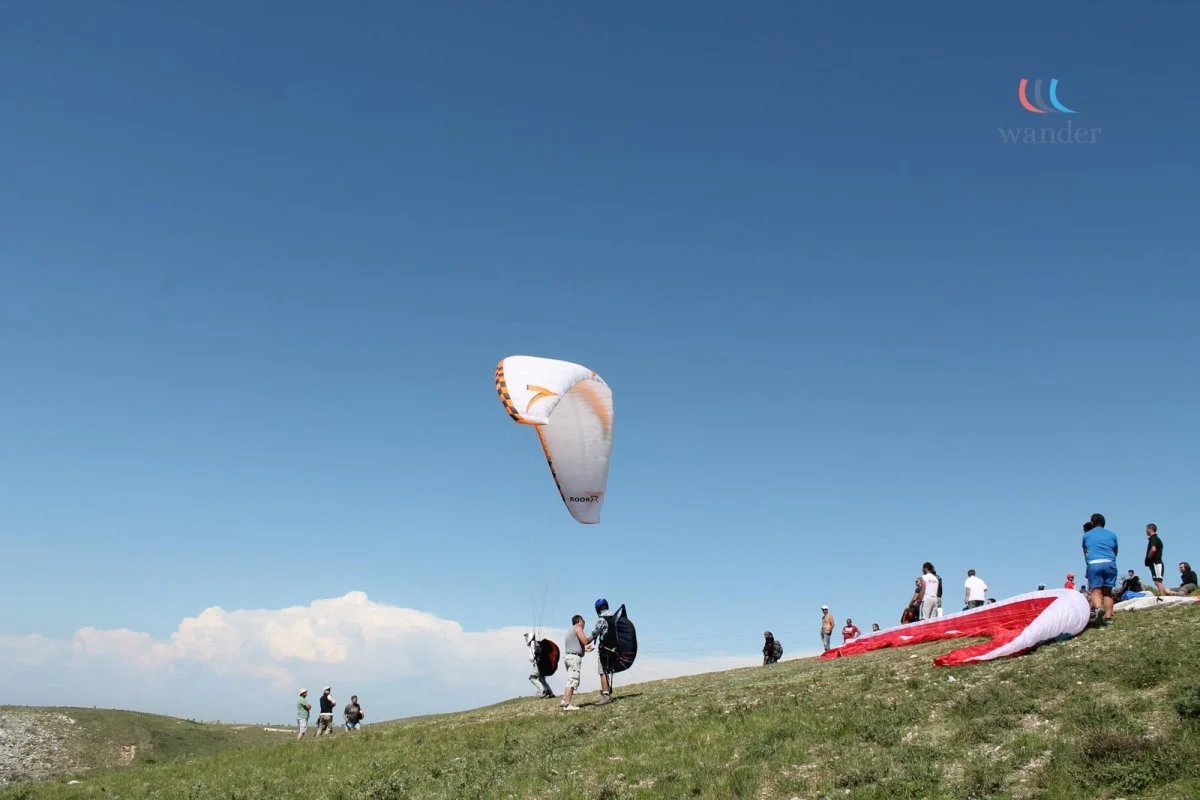 People preparing for a paragliding flight on a grassy hill under a clear blue sky.