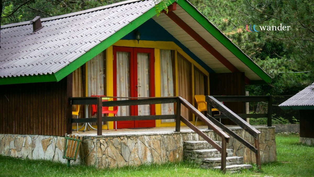 Colorful cabin with red, yellow, and blue accents on the door and window trim, set in a lush green environment with grass and trees, featuring a stone foundation, stairway, and small outdoor seating area.