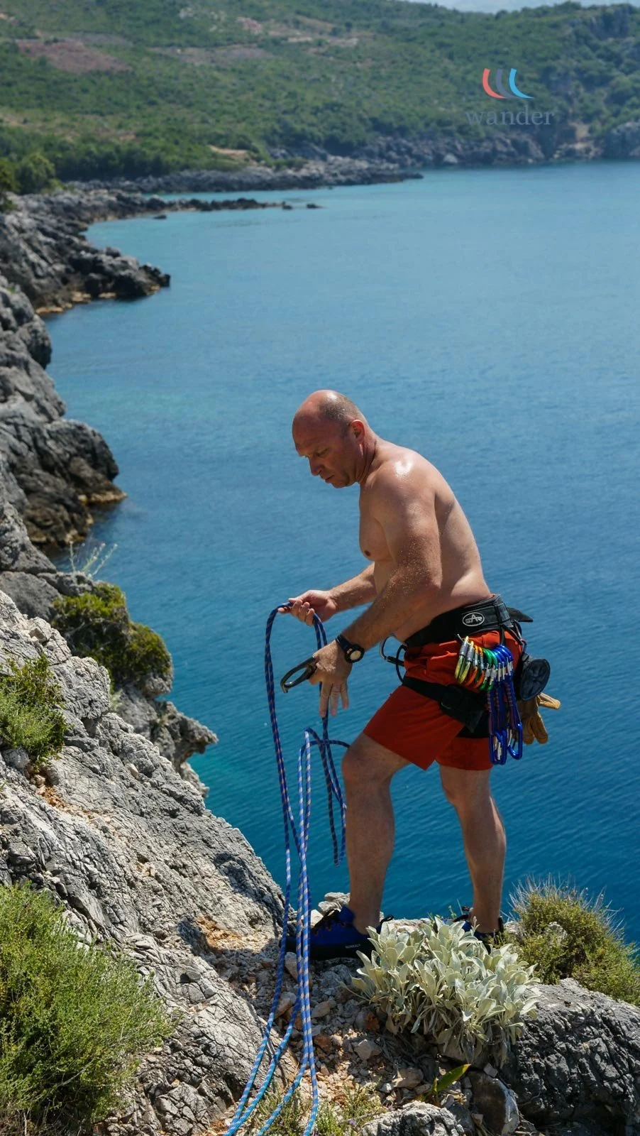A shirtless man with climbing gear stands on rocky terrain near water, preparing a rope across a cliff edge overlooking a blue bay and green hills.