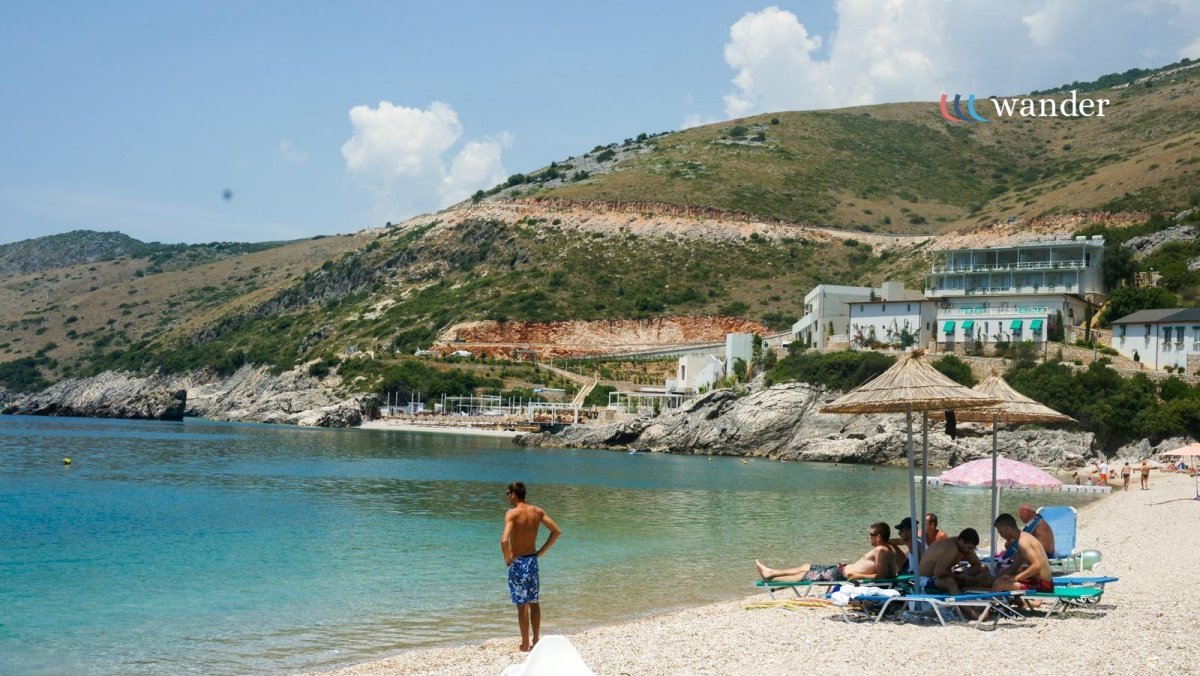 People relaxing on lounge chairs under umbrellas on a sandy beach with a scenic hillside in the background.