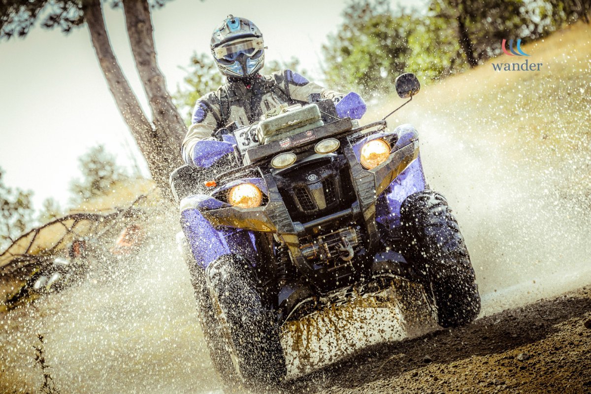 Person riding a blue all-terrain vehicle (ATV) through a muddy trail with water spray, wearing a helmet and riding gear, in a wooded outdoor setting.