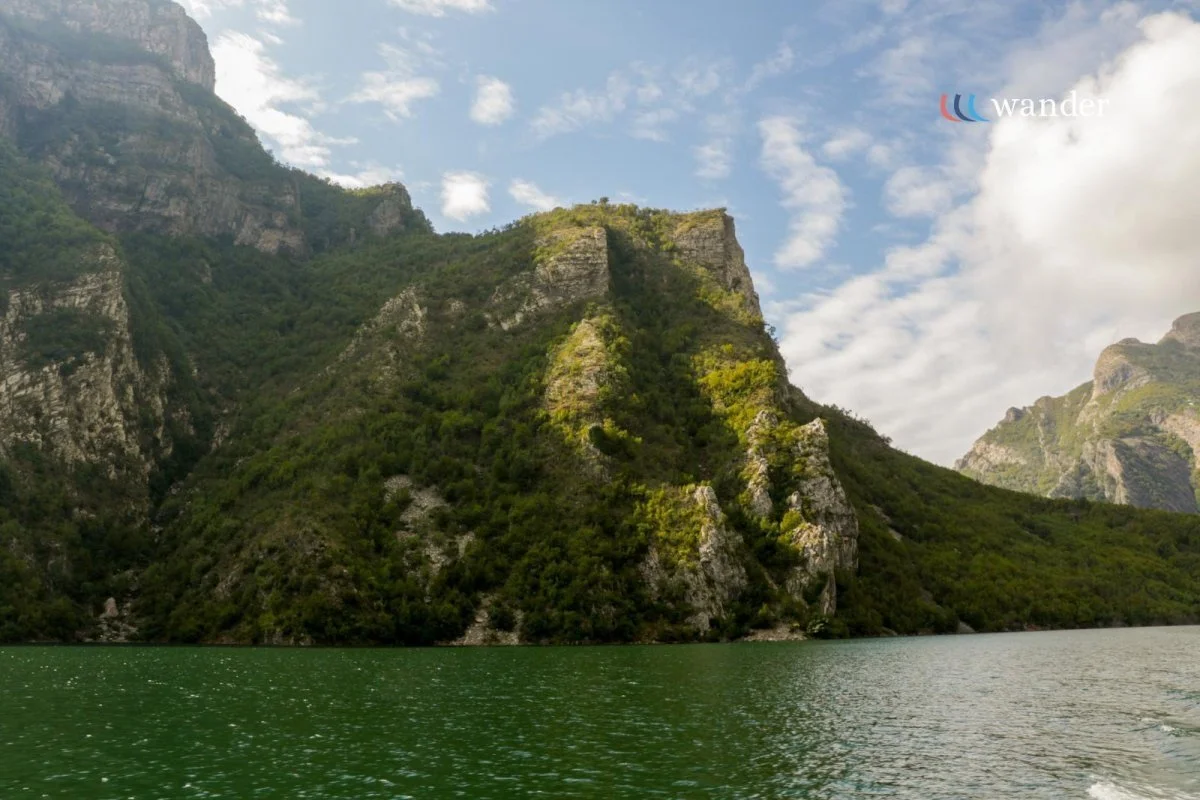 Scenic view of a green lake surrounded by tall, rugged mountains with some trees and bushes, under a partly cloudy sky.