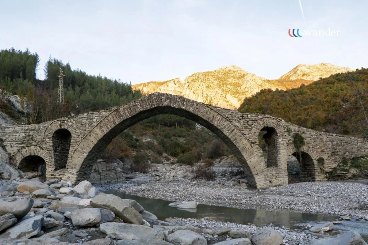 An ancient stone arch bridge over a rocky river, surrounded by mountains and forested hills.