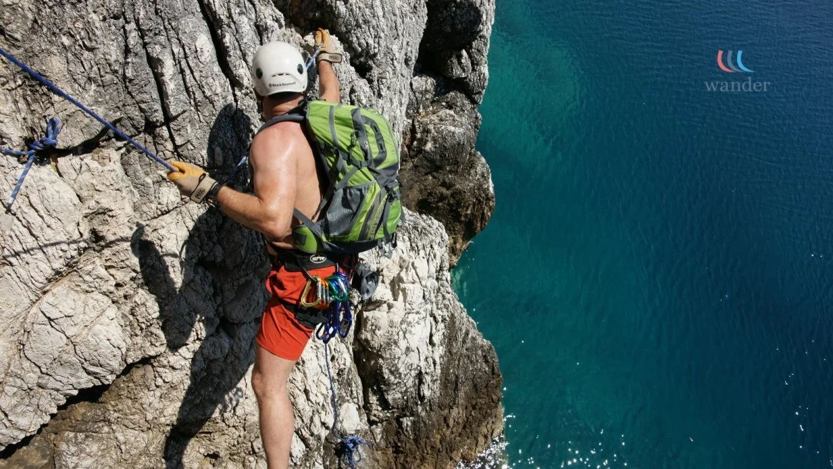 A man rock climbing on a steep cliff overlooking the water, wearing a helmet, harness, and backpack.