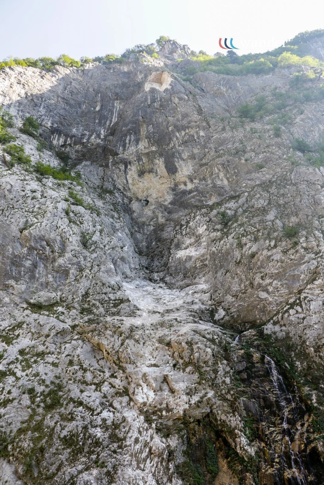 Tall rocky mountain with patches of greenery, a waterfall flowing down the cliff, and a partly cloudy sky.