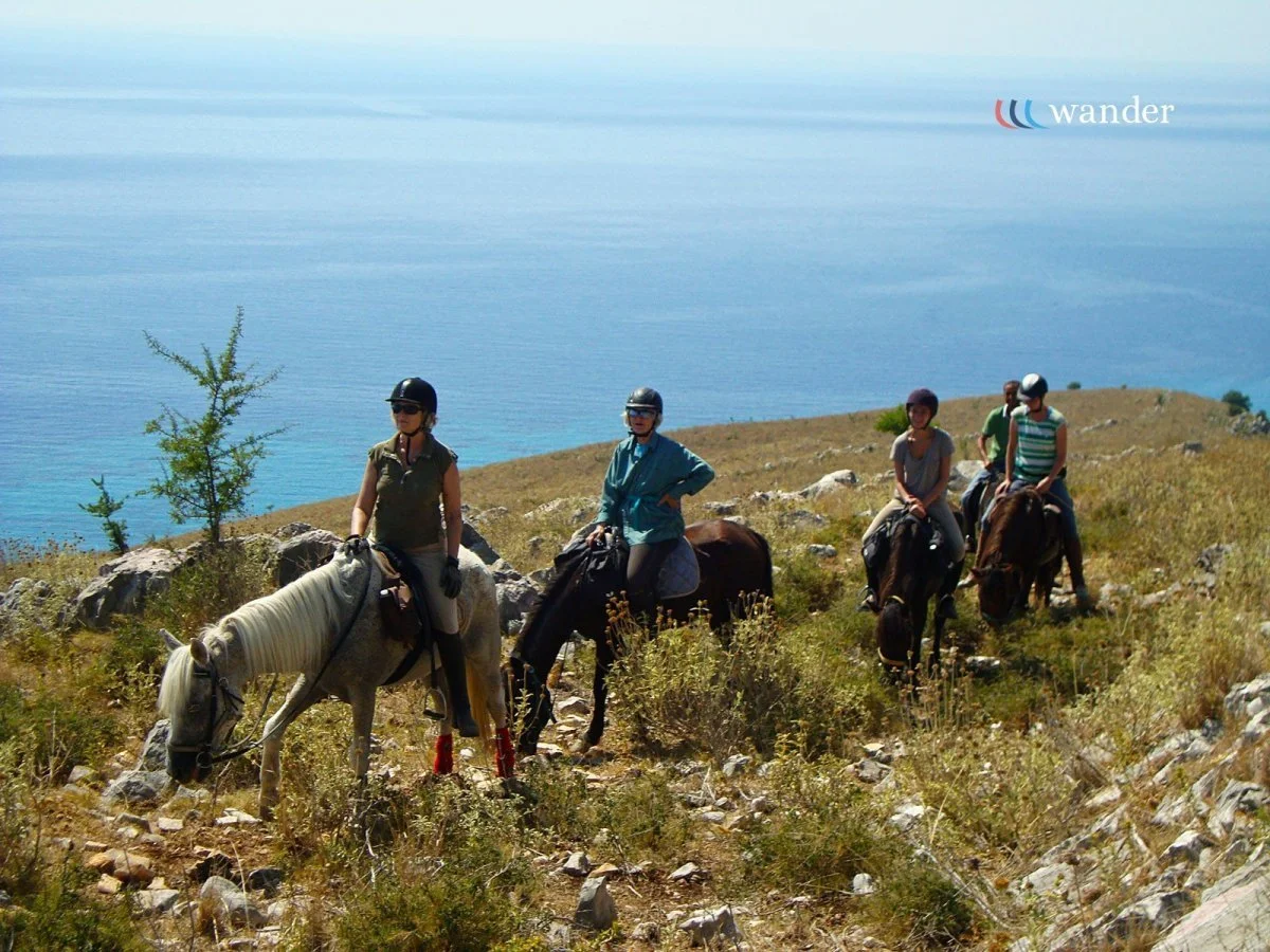 Four people riding horses along a rocky trail on a hillside near a body of water, with one woman in the front, two women in the middle, and two men in the back, wearing helmets and casual clothing.