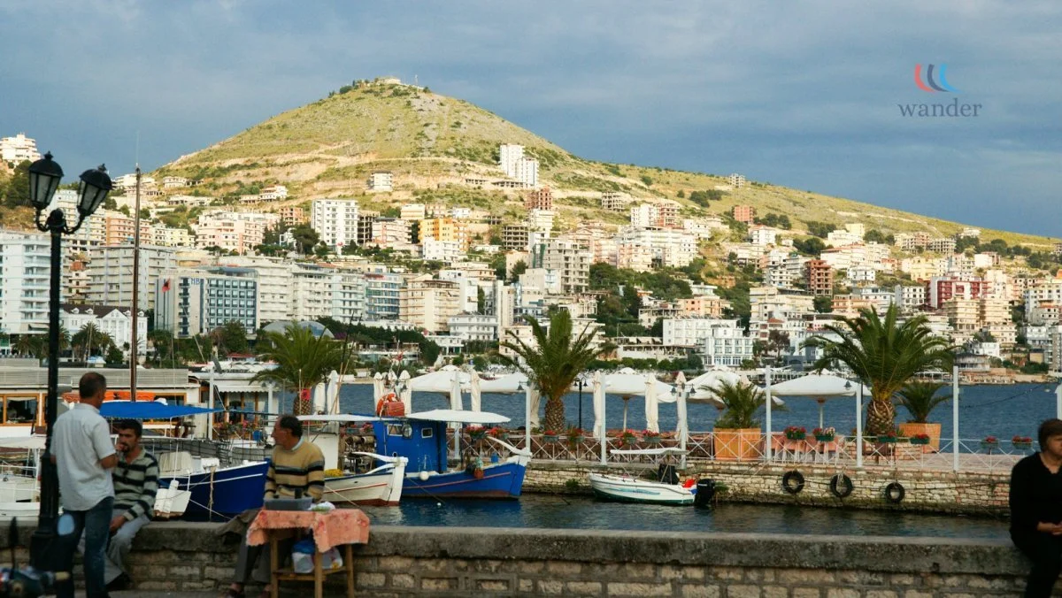 A waterfront scene with boats docked at a marina, palm trees, and people relaxing. In the background, there is a hillside city with numerous buildings and a mountain peak under a cloudy sky.