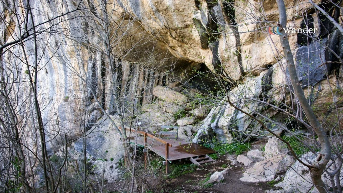 A trail with a small wooden footbridge in a rocky forest area with bare trees and a cliff face overhead.