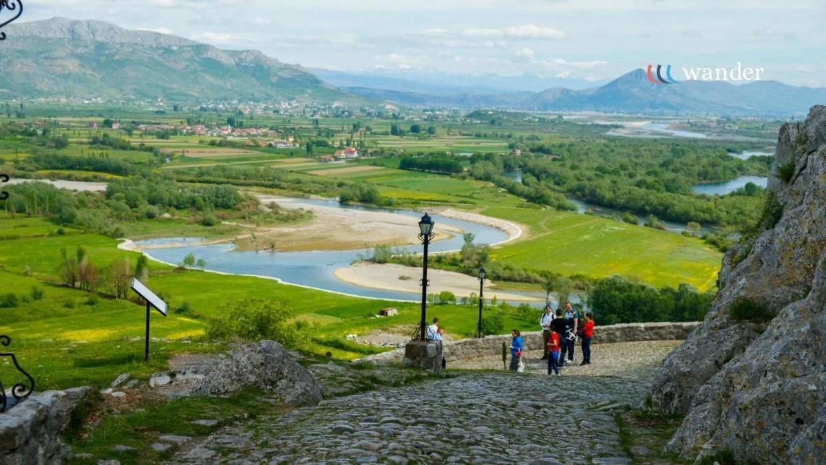 People viewing a scenic landscape with a river, green fields, mountains, and a cloudy sky, seen from a hilltop with cobblestone path and lamps.