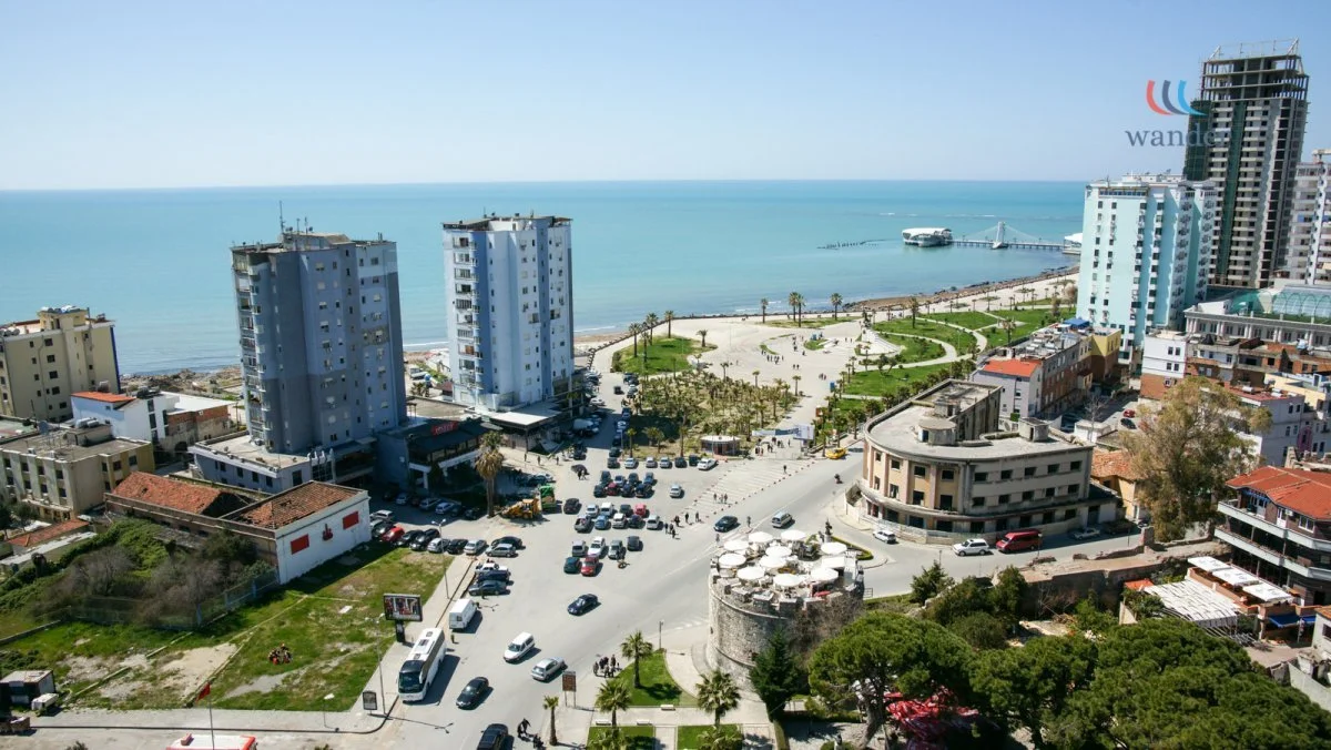 A coastal cityscape with high-rise buildings near the ocean, a park area with palm trees, and a beachfront with a pier and boat.