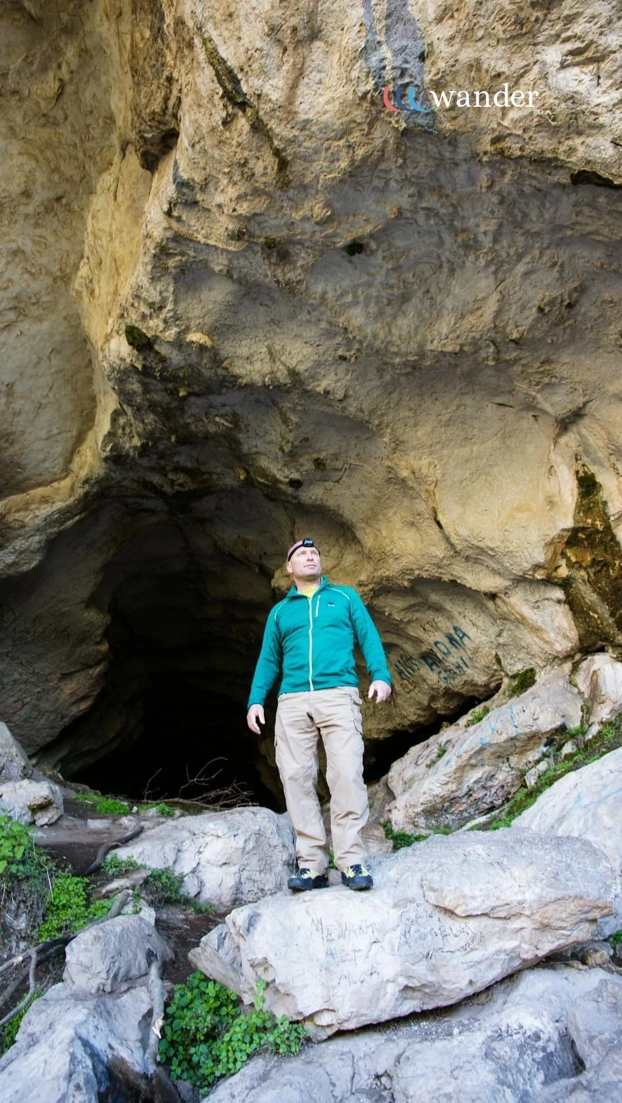 A man stands at the entrance of a large cave, wearing outdoor clothing and a headlamp, with graffiti on the cave walls and rocks around him.