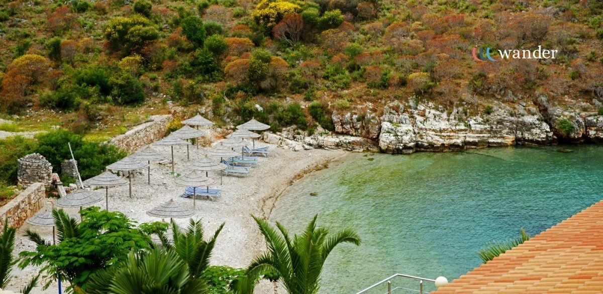 A secluded beach with lounge chairs and umbrellas, surrounded by greenery and rocky cliffs, with calm green water in a cove.