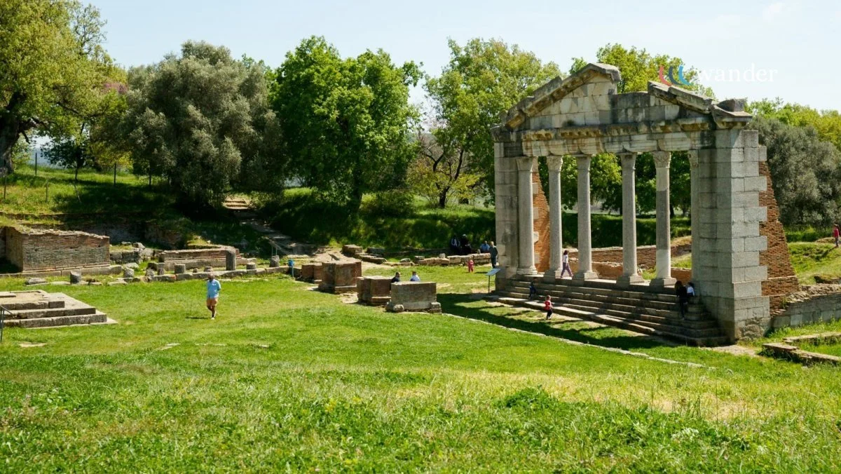 Ancient ruins with a stone temple featuring tall columns and stairs, surrounded by green grass and trees, with visitors exploring the site.