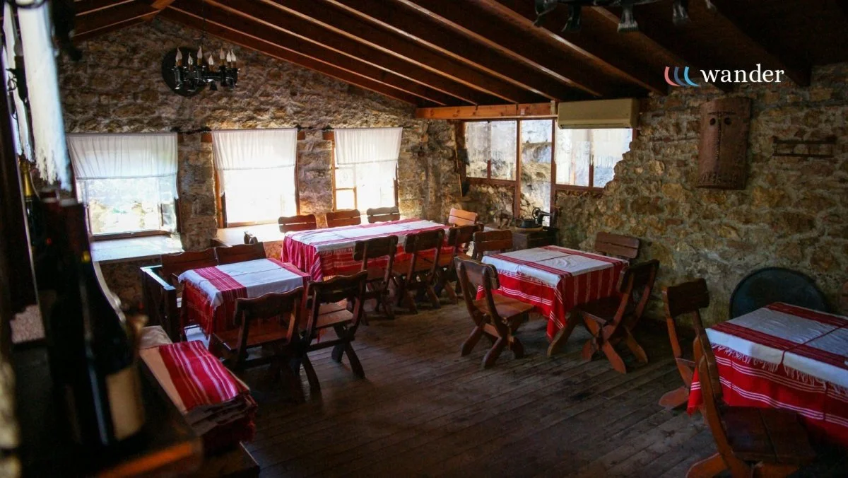 A rustic dining room with stone walls, wooden beams on the ceiling, and several tables covered with red and white striped tablecloths. There are multiple wooden chairs around each table, and windows with white curtains allowing natural light to fill the space.